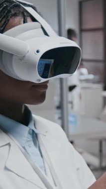 Sanal gerçeklik kulaklığı takan, dijital tablet üzerinde çalışan, ciddi hastalıkların tedavisini geliştirmek için yüksek teknoloji kullanan kadın koyu tenli tıp bilimcisinin dikey yan görüntüsü.