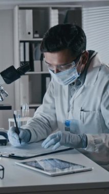 Koruyucu eldivenler ve yüz maskesi takmış, mikroskop altında mikroorganizmaları inceleyen ve modern laboratuarda notlar alan erkek tıp uzmanının dikey yavaş çekimi.