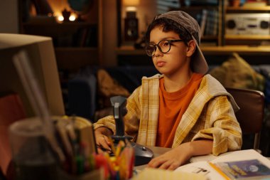 Gözlük ve kulaklık takan genç bir çocuk, arka planda kitaplarla ve elektronik cihazlarla dolu rahat çalışma odasında bilgisayar kullanırken masada oturuyor.