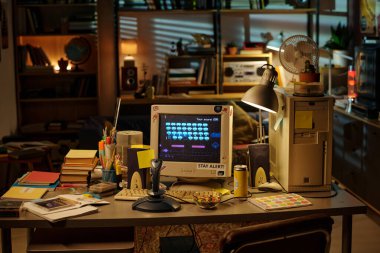 Eski bir bilgisayar ekranı ve video oyunu içeren Retro temalı çalışma alanı. Masa kitaplar, kağıtlar ve kırtasiye malzemeleriyle dolu. Ofis ortamında nostaljik bir ortam yaratıyor.