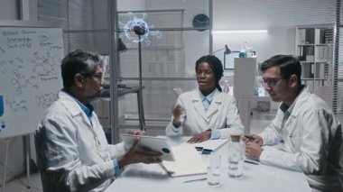 Üç farklı meslektaşın modern laboratuvarda buluşup virüs tedavisini tartışırken, arka planda 3 boyutlu hologram fanının kurşun virüs sergilediği orta ölçekli bir fotoğraf.