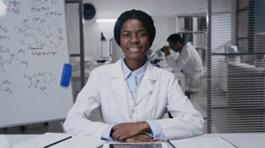 Laboratuardaki masada poz veren ve arka planda kimyasal bileşikler bulunan beyaz tahtaya mutlu bir şekilde bakan önlüklü mutlu Afrikalı Amerikalı tıbbi araştırmacının orta yavaş portresi.