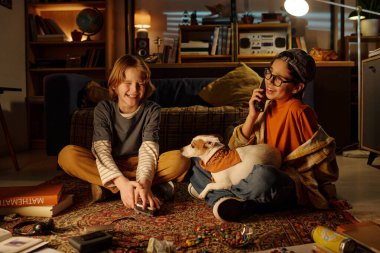 İki çocuk yerde oturmuş gülümsüyor ve evcil köpekle oynuyor. Ilık aydınlatma ve gündelik ortam, arka planda çeşitli nesnelerin dağıldığı rahat bir atmosfer yaratır.