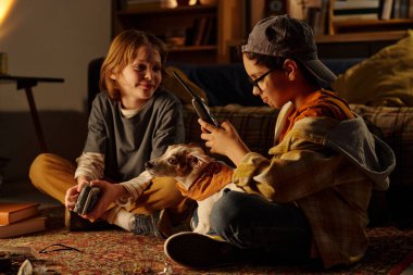 Genç arkadaşlar yerde oturuyor ve akşamın tadını çıkarıyorlar. Cihazları kullanıyorlar ve oturma odasında köpekle oynuyorlar. Etrafında çeşitli eşyalar var.