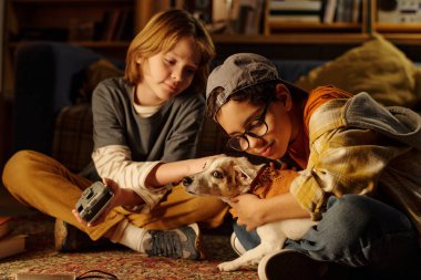 İki çocuk, sıcak ışıklandırma, günlük kıyafetler ve rahat bir atmosfer yaratan ev dekorasyonu içeren rahat bir ortamda küçük köpekle oynamaktan zevk alıyor.