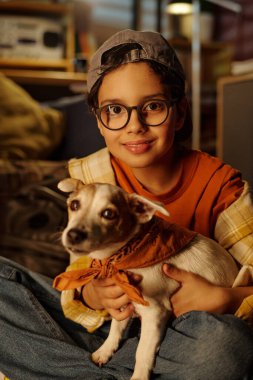 Çocuk sandalyede oturuyor ve gülümsüyor. Köpeği boynunda turuncu eşarpla tutuyor. Sıcak ışıklandırmalı rahat bir odada.
