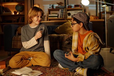 İki çocuk, arka planda kitap rafları ve kürenin olduğu sıcak aydınlatılmış oturma odasındaki desenli halıda kağıt oynamaktan zevk alıyor.