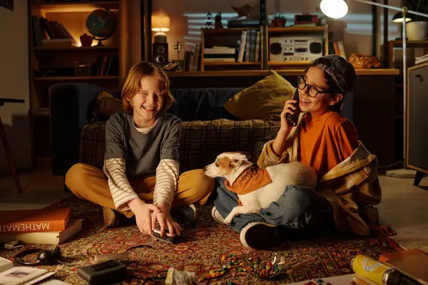 İki çocuk yerde oturmuş gülümsüyor ve evcil köpekle oynuyor. Ilık aydınlatma ve gündelik ortam, arka planda çeşitli nesnelerin dağıldığı rahat bir atmosfer yaratır.