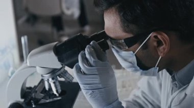 Koruyucu gözlüklü, yüz maskeli ve eldivenli erkek laboratuvar asistanının mikroskop altında numune incelerken uyuşturucu geliştirme laboratuarında deneyler yaparken yavaş çekim görüntüsü.