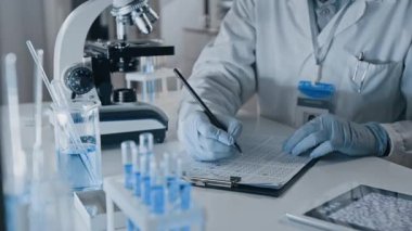 Modern ilaç geliştirme laboratuvarında mikroskop altında mikroorganizmaları incelerken koruyucu yüz maskesi, gözlük ve eldiven takan erkek tıp bilimcilerinin yan görüntüleri.