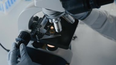 Laboratuvardaki mikroorganizmaların tıbbi araştırmaları sırasında objektif lensleri mikroskopta değiştiren isimsiz bir bilim adamının eldivenli ellerine yakın çekim.