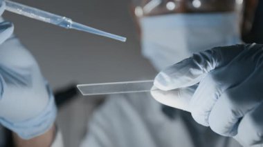 Koruyucu maskeli bilim adamının yakın plan çekimi cam kaydırağa bir damla çözelti ekleyip tıp laboratuarında mikroskop altında incelerken.