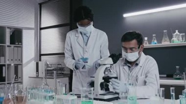 Erkek tıp bilimcisinin mikroskop altında numune incelerken ve kadın asistanın test tüpünde çözelti hazırlarken, modern laboratuarda çalışırken, koruyucu gözlük ve maske takarken orta ölçekli bir fotoğrafı.