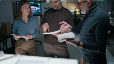 Üç beyaz erkek ve kadın araba tasarımcısının, yeni çapraz model modifikasyonu, taslakları, çizimleri ve ölçekli modelleri gözden geçirme ve dış tasarım hakkındaki tartışmaları tartıştığı orta ölçekli bir fotoğraf.