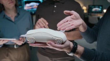 Orta yaşlı erkek araba mühendisinin orta ölçekli yakın çekimi elinde küçük çaplı yeni bir geçiş modeli, dış özelliklere işaret ederken aynı zamanda ofisteki takım arkadaşlarını ikna ediyor.