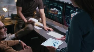 Otomotiv Tasarım Bürosu 'ndaki kadın ve erkek araba mühendislerinin, mevcut projeleri tartışırken bilgisayar ve tabletteki otomobil çizimlerini izledikleri orta büyüklükte bir fotoğraf.