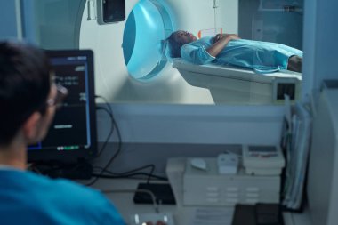 MRI makinesinin içinde yatan hasta kontrol odasından tıbbi uzman monitörleri izliyor. Oda gelişmiş tıbbi ekipman ve vitrinlerle dolu.