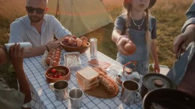 Küçük kız yaz kampı gezisi sırasında piknik masasında aile yemeği hazırlarken annesine portatif fırında pastırma ve yumurta pişirmesi için yardım ediyor.