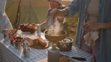 Anne taşınabilir fırında yemek hazırlarken çocuklar ve kocalar güneşli bir günde kamp alanında piknik masasında sohbet ediyorlar.