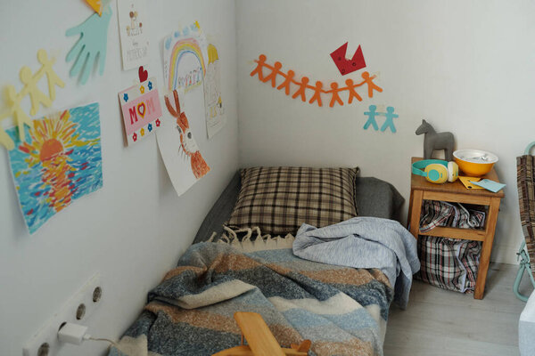 Children drawings hanging on wall above sleeper of refugee with pillow and plaid standing in the corner of crisis camp