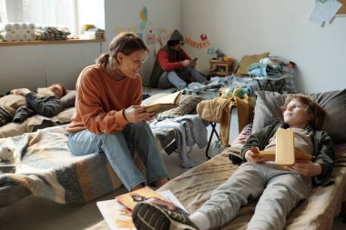 Elinde açık bir kitapla uyuyan oğluna bakıp mülteci kampında hikaye dinleyen mutlu genç bir anne.