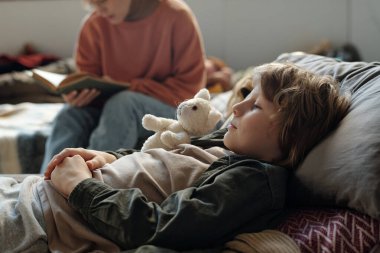 Beyaz yumuşak oyuncaklı şirin bir çocuğun başını yastığa koyup annesine karşı uyuyanların yanında kitap okurken dinlenmesi.