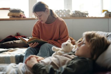 Mülteci kampında oğlunun önünde uyuyan küçük oğluna açık kitap okuyan bekar bir anneye odaklan.