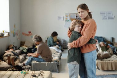 Genç oğlunu kucaklayan ve mülteci kampında dikilirken kameraya bakan sefil, genç bir kadın.