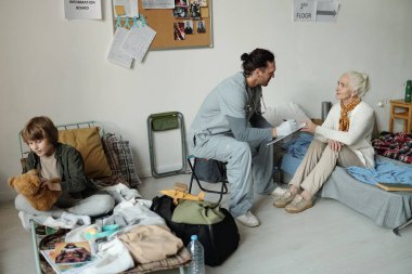 Tıbbi önlüklü genç adam, yaşlı kadın mültecilerin önünde oturup sorularını cevaplarken kâğıt belgelere not alıyor.