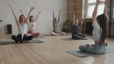 Sağlıklı kızlar ve kadın koçun eşliğinde yoga yapan Down sendromlu genç bir kadının yaklaşım yavaşlaması. Parlak stüdyoda yoga minderlerinde birlikte oturuyorlar. Engellilere eşit haklar kavramı