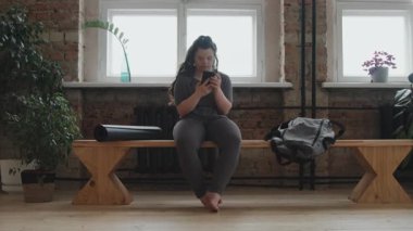 Ağır ilerleyen sendromlu genç kadın akıllı telefon kullanarak modern yoga dersinde pencerenin önündeki bankta oturuyor.