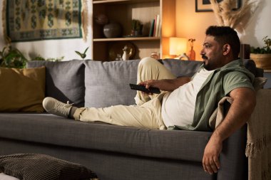 Sıcak oturma odasındaki kanepede uzanan adam, uzaktan kumandayı tutuyor ve arkasını yaslıyor, sıcak ışıklandırma ve dekor elementleriyle çevrili.