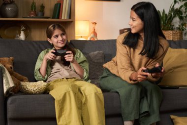 Genç kız ve kadın kanepede oturuyor, her biri oyun kumandalarını tutuyor ve video oyunları oynarken neşeli anları paylaşıyorlar, yanlarında patlamış mısır kasesi ve rahat oda ayarlarıyla.