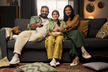 Üç kişilik bir aile kanepede oturur, film izler ve patlamış mısır yer. Rahat oturma odası, yastıklar, battaniyeler ve sıcak ışıklandırma.