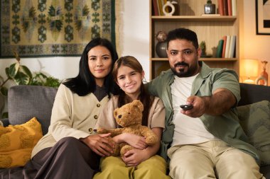 Çok ırklı bir aile, oturma odasındaki kanepede rahat bir oyuncak ayı tutarak rahat vakit geçiriyorlar. Ebeveynler televizyon izlerken gülümseyen çocuğu kucaklıyor