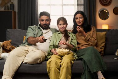 Üç kişilik aile rahat bir koltukta oturup film seyrediyor ve atıştırmalıkların tadını çıkarıyor.