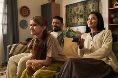 Aile, içi doldurulmuş oyuncaklarla kanepede oturup kaliteli vakit geçirmenin keyfini çıkarıyor. Arkaplanda dekoratif öğelerle sıcak ve davetkar ortam