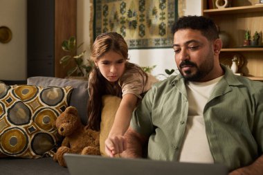 Orta yaşlı bir adam kızına ev ödevlerinde yardım ediyor rahat oturma odasında dekoratif yastıklar, oyuncak ayı ve duvar örtüsü ile olay yerine sıcaklık katıyor.