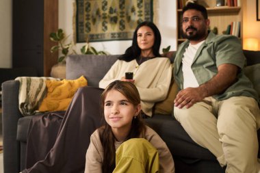 Üç kişilik çok ırklı aile kanepede oturmuş bitki ve sanat eserleriyle süslenmiş rahat oturma odasında televizyon seyrediyorlar.