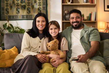 Gülümseyen aile portresi rahat bir kanepede oturmuş oyuncak ayı tutuyor ve evde güzel vakit geçirip dinleniyor.