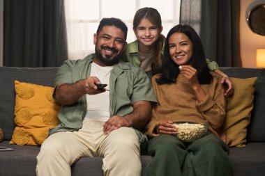 Aile, patlamış mısırları paylaşırken ve gülümserken kanepede rahatlarken eğleniyor. Kızı anne ve babasına sarılıyor, rahat bir yaşam alanında mutluluğu gösteriyor.