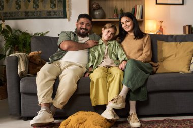 Üç kişilik aile sıcak bir oturma odasında kanepede dinleniyor. Ilık ışıklar ve duvardaki dekore edilmiş raflar sıcak bir ortam yaratıyor.