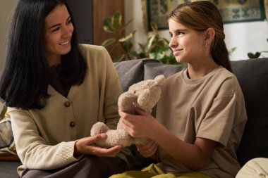 Gülümseyen İspanyol anne kanepeye oturmuş kızıyla birlikte oyuncak ayıyı tutuyor birbirlerine yaslanıyor ve evlerinde bitkilerle çevrili samimi anları paylaşıyorlar.