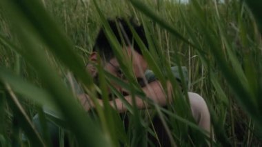 Gözlüklü, sıradan haki gömlekli, sakallı, yeşil çimlerin arkasından kameraya bakan yakışıklı Afrikalı genç bir adamın orta boy portresi.