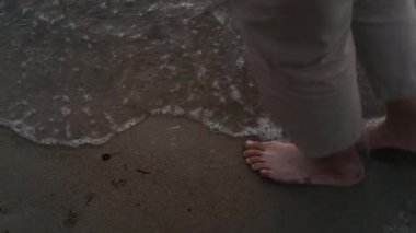 Ortalama yakın çekim ardından haki pantolonlu, tanınmayan bir adamın ayakları ve bacakları kumlu okyanus sahillerinde dalgalar halinde yürürken, akşam dışarıda tek başına dinlenirken.