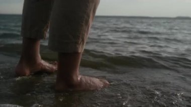 Bej pantolonlu, gölde ya da deniz sahilinde kumların üzerinde yürüyen, çıplak tenli, serin su hissini hisseden ve zevk alan isimsiz bir adamın ayaklarına yakın dur.