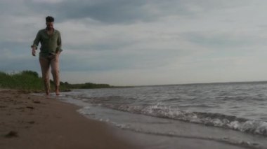 Dalgın genç Afrikalı Amerikalı adamın kumlu deniz ya da göl sahillerinde yürürken, serin dalgaların tadını çıkarırken, akşam vakti dışarıda yalnız başına eğlenirken geniş ve yavaş bir çekim.