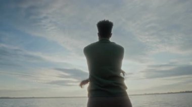 Gölün kıyısında duran yakışıklı Afrikalı Amerikalı adamın orta boy arka portresi. Suyun üzerinde gün batımını seyrediyor, kollarını sallıyor, sonra kameraya bakıyor, gülümsüyor.
