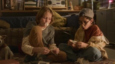 İki gencin sakızlarıyla baloncuk çıkardığı ve loş ışıklandırılmış ev ortamındaki araba görüntülerine baktığı bir kare.