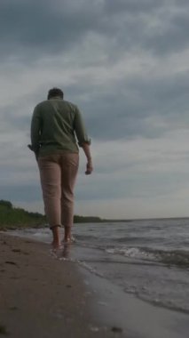 Haki gömlek ve pantolon giyen, kumlu deniz veya göl sahillerinde çıplak ayakla yürüyen ve akşam vakti doğada tek başına dinlenen tanınmamış bir adamın dikey arka çekimi.